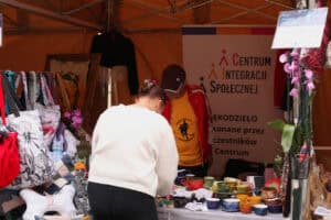 Dwie osoby obsługują stoisko targowe. Za nimi znajduje się baner „Centrum Integracji Społecznej” i regał z materiałowymi torbami.