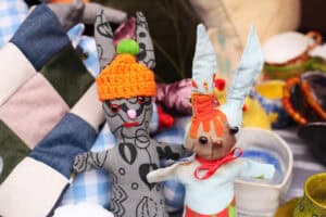 Dwie ręcznie szyte maskotki królików siedzą na stosie poduszek. Królik po lewej ma szary wzorzysty materiał i pomarańczową czapkę. Królik po prawej ma jasne kolory i czerwoną kokardkę.