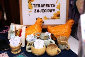 Na stole stoi ramka z napisem „Terapeuta zajęciowy”. Przed ramką stoją dwie pomarańczowe ceramiczne figurki kur, małe gliniane kubeczki i suszony bukiet.