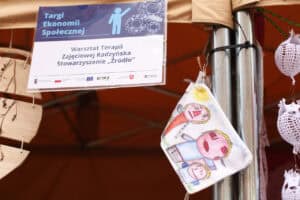 Na stoisku wisi płócienna torba z dziecięcym rysunkiem trzech postaci. Obok znajduje się fragment baneru z napisem „Warsztat Terapii Zajęciowej Radzyńska, Stowarzyszenie „Źródło””.