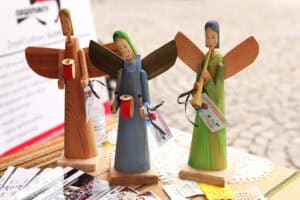 Na stole stoją trzy ręcznie rzeźbione i malowane drewniane figurki aniołów. Środkowy anioł jest niebieski i trzyma bębenek. Pozostałe figurki są zielona i mają kolor naturalnego drewna.