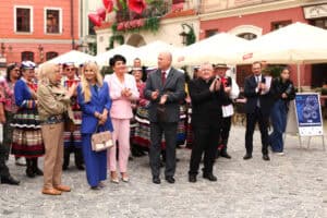 Grupa osób stoi na brukowanym placu podczas wydarzenia plenerowego. Wśród nich są Anna Augustyniak i ks. prałat Tadeusz Pajurek.
