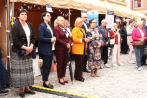 Grupa osób stoi w rzędzie przy zadaszonych stoiskach targowych. Wśród nich są Joanna Olszewska i Monika Wac. Uczestnicy obserwują wydarzenie plenerowe.