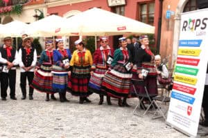 Grupa kobiet i mężczyzn w tradycyjnych, kolorowych strojach ludowych stoi na brukowanym placu obok baneru informacyjnego ROPS Lublin.