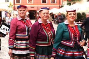 Trzy kobiety w tradycyjnych, kolorowych strojach ludowych i czepkach stoją blisko siebie. Wokół znajdują się zabytkowe kamienice i białe parasole.