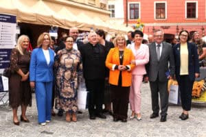 Około dziesięciu dorosłych osób stoi w rzędzie na brukowanym placu i pozuje do zdjęcia. Za nimi znajdują się stoiska targowe.