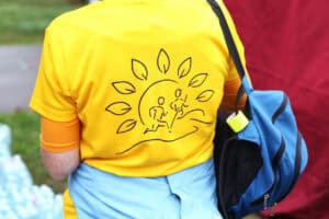 Plecy uczestnika biegu w żółtej koszulce z logo wydarzenia: dwie biegnące sylwetki ludzi na tle grafiki wschodzącego słońca z promieniami w kształcie liście.