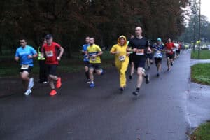 Grupa biegaczy biegnie asfaltową ścieżką. Wśród nich jest uczestniczka w żółtym kombinezonie z kapturem przypominającym strój maskotki.