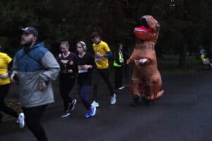 Uczestnicy biegną parkową aleją. Wśród nich jest osoba w dmuchanym kostiumie dinozaura T-Rexa.