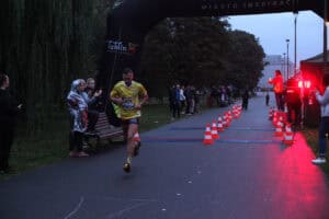 Biegacz w żółtej koszulce z numerem 309 przekracza metę pod czarną bramą z napisem „Lublin Miasto Inspiracji”. Obok znajduje się podświetlony na czerwono zegar i pomarańczowe pachołki.