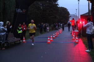 Biegacz w żółtej koszulce z numerem 266 przekracza metę wyznaczoną pachołkami. Po bokach stoją kibice, a obok znajduje się elektroniczny zegar wyników.