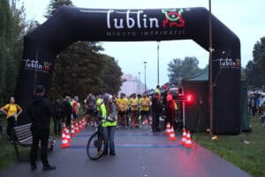 Duża czarna dmuchana brama startowa z napisem „Lublin Miasto Inspiracji” stoi nad parkową aleją. Przy starcie gromadzą się uczestnicy biegu.