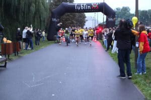 Grupa biegaczy rusza spod czarnej dmuchanej bramy z napisem „Lublin Miasto Inspiracji”. Po obu stronach alei stoją kibice i obsługa wydarzenia.
