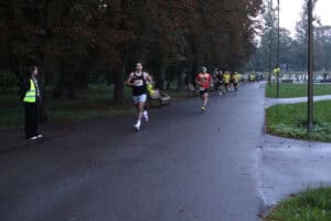 Uczestnicy biegną parkową aleją. Na pierwszym planie jest mężczyzna w czarnej koszulce bez rękawów. W tle osoba w odblaskowej kamizelce zabezpiecza trasę.