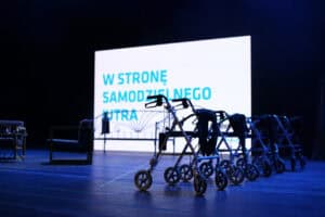 Na scenie teatralnej oświetlonej niebieskim światłem stoi rząd chodzików rehabilitacyjnych. W tle biały ekran z napisem „W stronę samodzielnego jutra”.