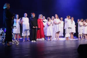 Aktorzy stoją w rzędzie na scenie w świetle reflektorów. Po lewej stronie stoi Maria Pietrusza-Budzyńska w czarnym stroju.
