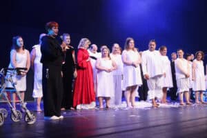 Maria Pietrusza-Budzyńska trzyma mikrofon i przemawia na scenie. Obok niej stoi liczna grupa aktorów w białych kostiumach.