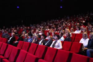 Publiczność siedzi na czerwonych fotelach w sali teatralnej i obserwuje scenę.
