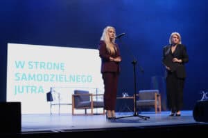 Anna Augustyniak przemawia do mikrofonu na scenie teatralnej. W tle telebim z napisem „W stronę samodzielnego jutra”.