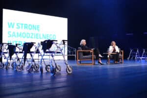 Debata na scenie teatralnej. Dwie kobiety siedzą w fotelach i rozmawiają. Z przodu stoją chodziki rehabilitacyjne. Z tyłu za nimi widać ekran z napisem „W stronę samodzielnego jutra”.