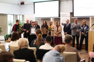 Grupa nagrodzonych osób i organizatorów stoi na scenie. Jedna z osób trzyma oprawiony certyfikat. Po prawej stronie mężczyzna przemawia do mikrofonu.