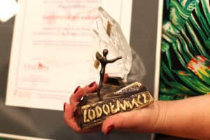 Dłonie trzymają statuetkę konkursu „Lodołamacze” z postacią ludzką na tle kry lodowej. W tle znajduje się fragment dyplomu.