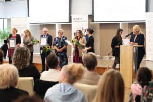 Grupa laureatów stoi na scenie z oprawionymi dyplomami i kwiatami. Laureaci są zwróceni w stronę publiczności podczas gali wręczenia nagród.