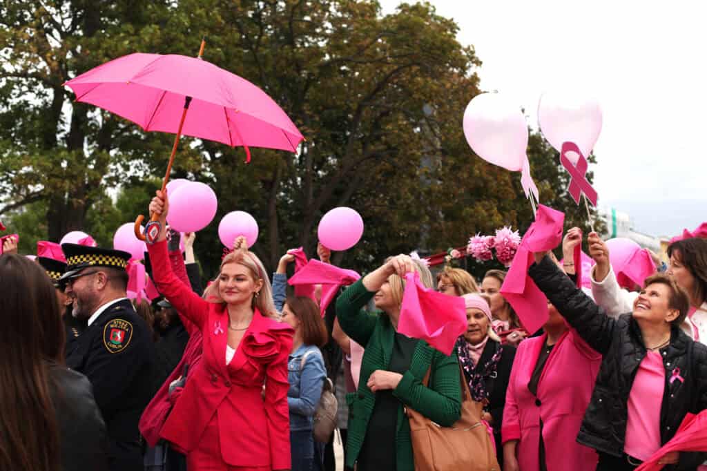 Kobieta trzyma wysoko otwarty różowy parasol. Inne uczestniczki marszu trzymają różowe chusteczki i balony. Wśród nich jest kobieta w zielonej marynarce. Obok stoi funkcjonariusz Straży Miejskiej.