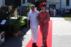 Para idzie czerwonym dywanem. Mężczyzna w koszuli w paski i białych spodniach podtrzymuje pod rękę kobietę w długiej czerwonej sukni i czerwonym fascynatorze z kwiatem.