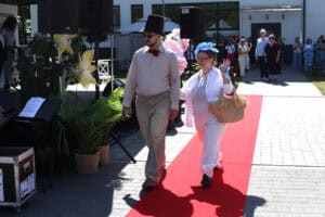Para idzie czerwonym dywanem. Kobieta w białym stroju i niebieskim fascynatorze macha do zgromadzonych, a obok niej idzie mężczyzna w jasnej koszuli i cylindrze.