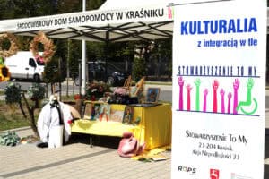 Stoisko plenerowe ŚDS w Kraśniku z ekspozycją obrazów malarskich oraz dekoracji artystycznych. Obok po prawej stronie stoi pionowy baner Stowarzyszenia „To My”.