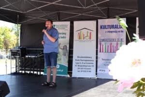 Na scenie stoi mężczyzna w niebieskiej koszuli i dżinsowych spodenkach i śpiewa do mikrofonu. W tle widać banery organizatorów wydarzenia.