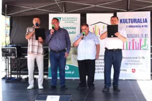Czterech mężczyzn stoi na scenie, śpiewa do mikrofonów i trzyma czarne teczki z tekstami. Wszyscy mają na sobie koszule i długie spodnie.