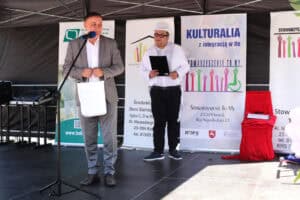 Mężczyzna w marynarce i szarych spodniach stoi przy mikrofonie na scenie i trzyma białą torbę. Za nim stoi konferansjer w białym smokingu i cylindrze z czarną podkładką w dłoni.