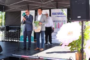Dwóch mężczyzn stoi na scenie przy mikrofonach i trzyma duży wiklinowy kosz z produktami. W tle stoi konferansjer w cylindrze.