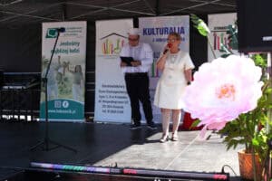 Agnieszka Niedziałek w jasnej sukni przemawia do mikrofonu na scenie plenerowej. Obok niej stoi konferansjer w białym garniturze i cylindrze z notesem w dłoni.
