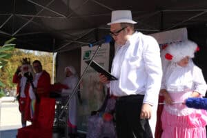 Konferansjer w białym smokingu i cylindrze stoi przy mikrofonie na scenie i czyta tekst z podkładki. W tle widać osoby w strojach z epoki.