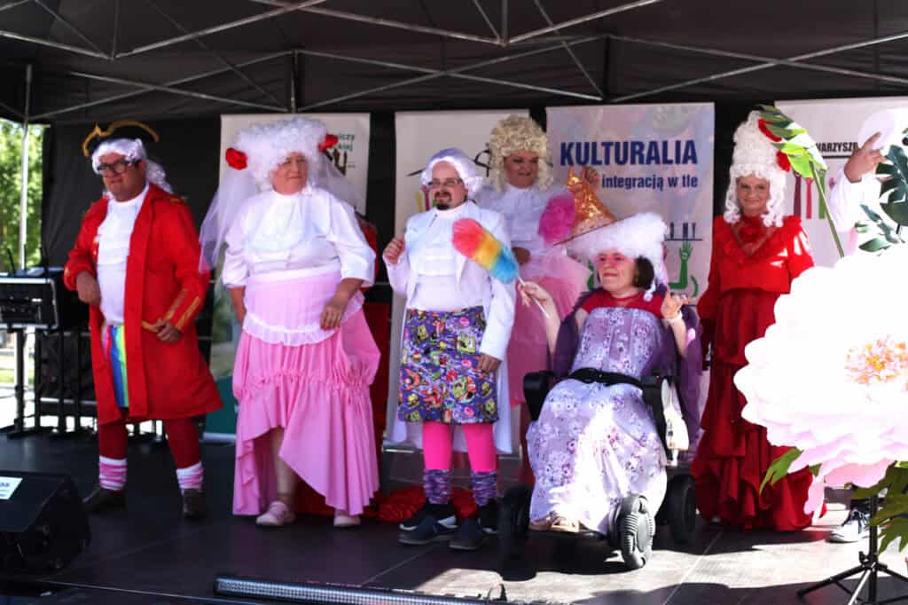 Grupa osób w barokowych strojach i perukach pozuje na scenie. Po prawej stronie uczestniczka w fioletowej sukni i kapeluszu z piórem siedzi na wózku.