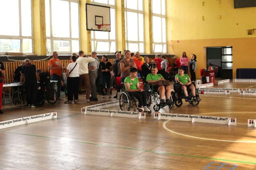 W hali sportowej podczas zawodów uczestnicy w zielonych koszulkach siedzą na wózkach przy boisku. W tle stoją kibice i organizatorzy. Wzdłuż boiska znajdują się niskie barierki z napisem „Integracyjne Centrum Sportu i Rehabilitacji Start”.
