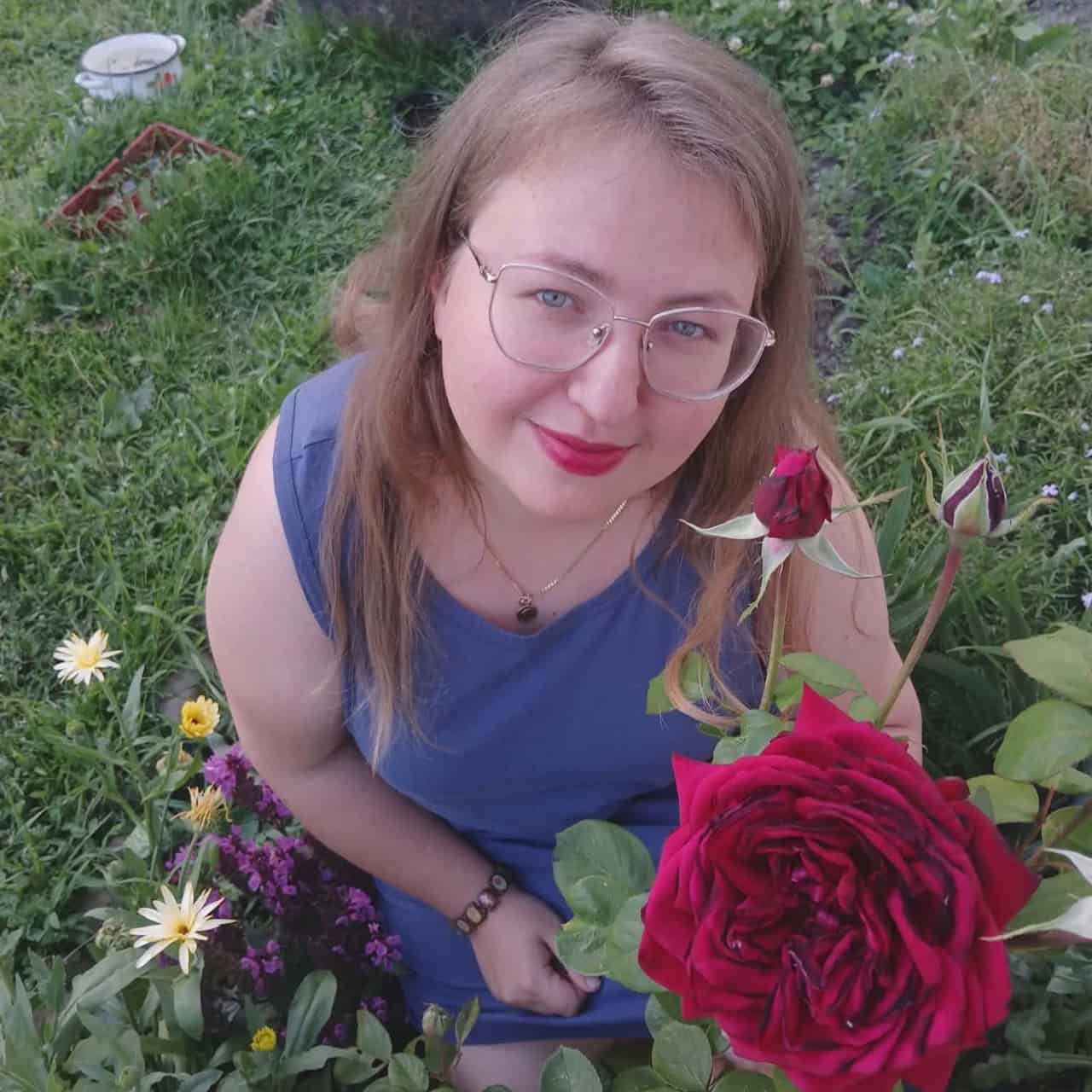 Katarzyna Wysokińska w plenerze pośród kwiatów, przy gałązce kwitnących bordowych róż.