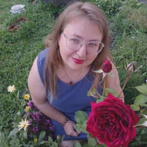 Katarzyna Wysokińska w plenerze pośród kwiatów, przy gałązce kwitnących bordowych róż.