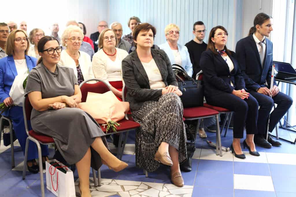 Uczestnicy spotkania siedzą w rzędach czerwonych krzeseł. Wśród nich są Monika Wac i Joanna Olszewska.