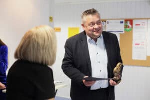 Uśmiechnięty Grzegorz Sołtys w ciemnym garniturze i błękitnej koszuli trzyma dyplom i mosiężną statuetkę. Obok stoi blondynka w czarnej sukience.
