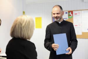 Uśmiechnięty ksiądz w czarnej sutannie trzyma niebieską teczkę. Obok stoi blondynka w czarnej sukience.