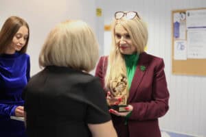 Anna Augustyniak trzyma mosiężną statuetkę w kształcie uścisku dłoni. Przed nią stoi kobieta w czarnej sukience.