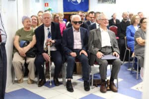 Cztery osoby siedzą obok siebie podczas uroczystości. Od lewej: kobieta w zielonej bluzce, starszy mężczyzna w garniturze z białą laską, mężczyzna w ciemnych okularach oraz uśmiechnięty mężczyzna w szarym garniturze.