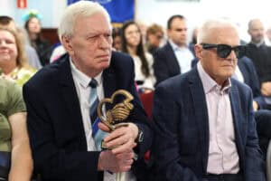 Dwaj mężczyźni siedzą obok siebie. Mężczyzna po lewej, w garniturze, trzyma mosiężną statuetkę w kształcie uścisku dłoni. Obok niego siedzi mężczyzna w ciemnych okularach.