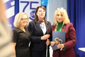 Trzy kobiety stoją na tle baneru „75 lat”. Od lewej: blondynka w czarnej sukience, Justyna Szafranek i Anna Augustyniak, która trzyma dyplom oraz pamiątkową statuetkę.