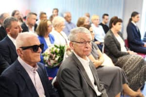 W pierwszym rzędzie sali konferencyjnej siedzi mężczyzna w ciemnych okularach przeciwsłonecznych oraz mężczyzna w przezroczystych okularach i szarym garniturze.