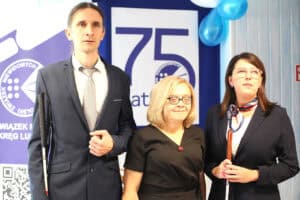 Trzy osoby stoją na tle baneru jubileuszowego z logo Polskiego Związku Niewidomych. Mężczyzna po lewej i kobieta po prawej trzymają białe laski.